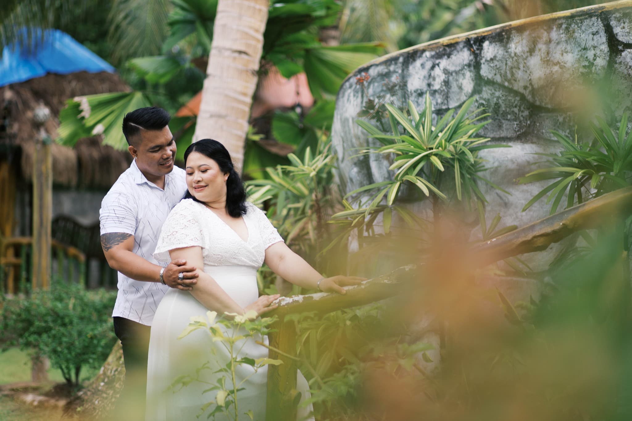 Smiling couple in white clothing poses affectionately in a lush, tropical outdoor garden setting.