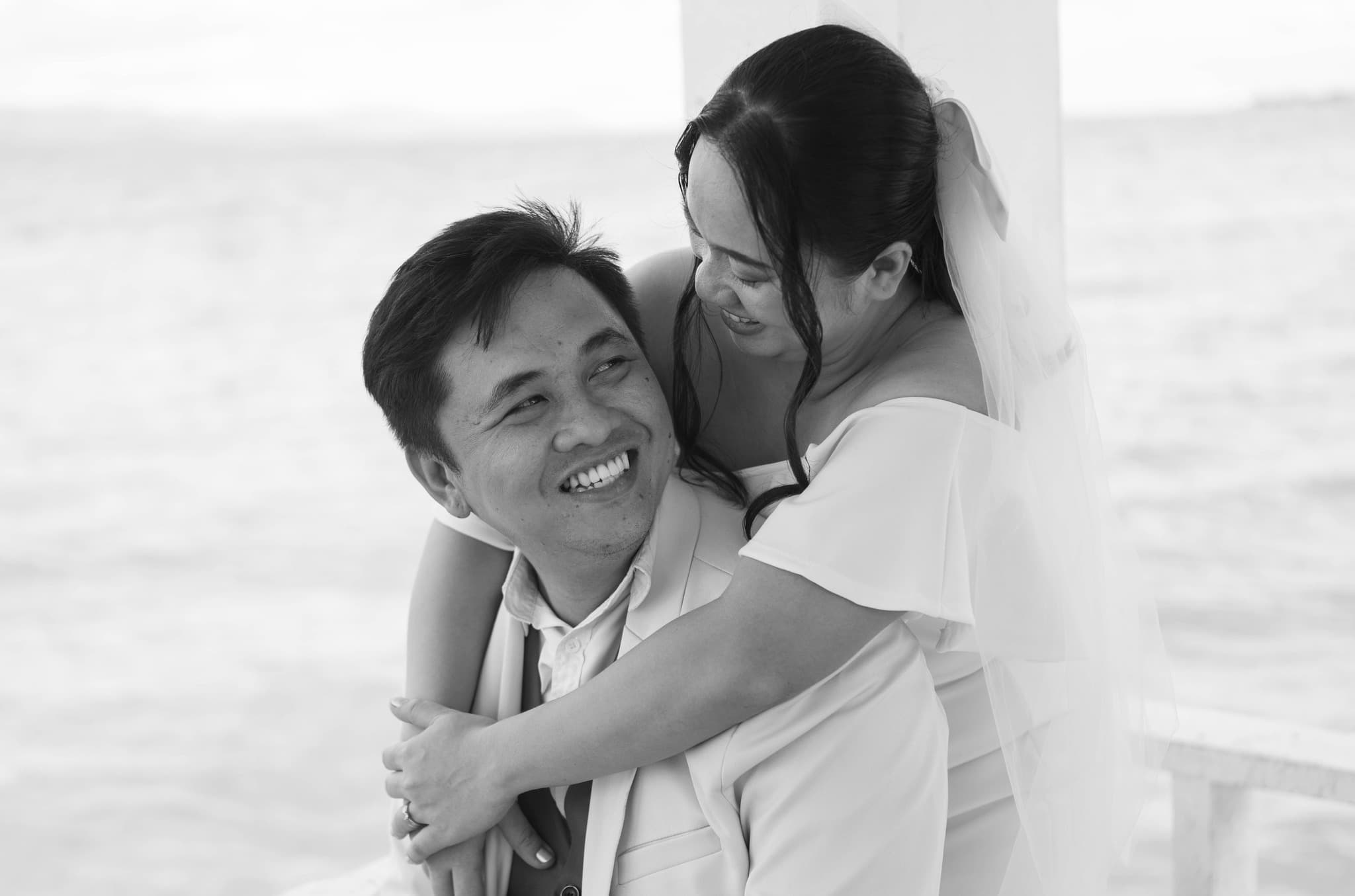 Smiling bride in a veil hugs her groom from behind in a monochrome seaside photo.