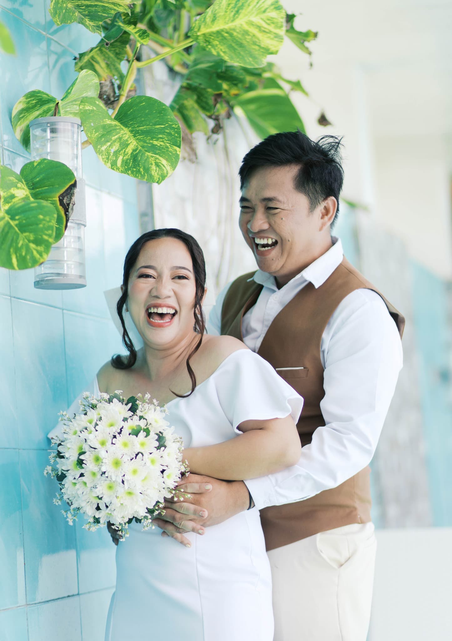 Joyful bride and groom laughing together against a blue tiled wall with green hanging plants.