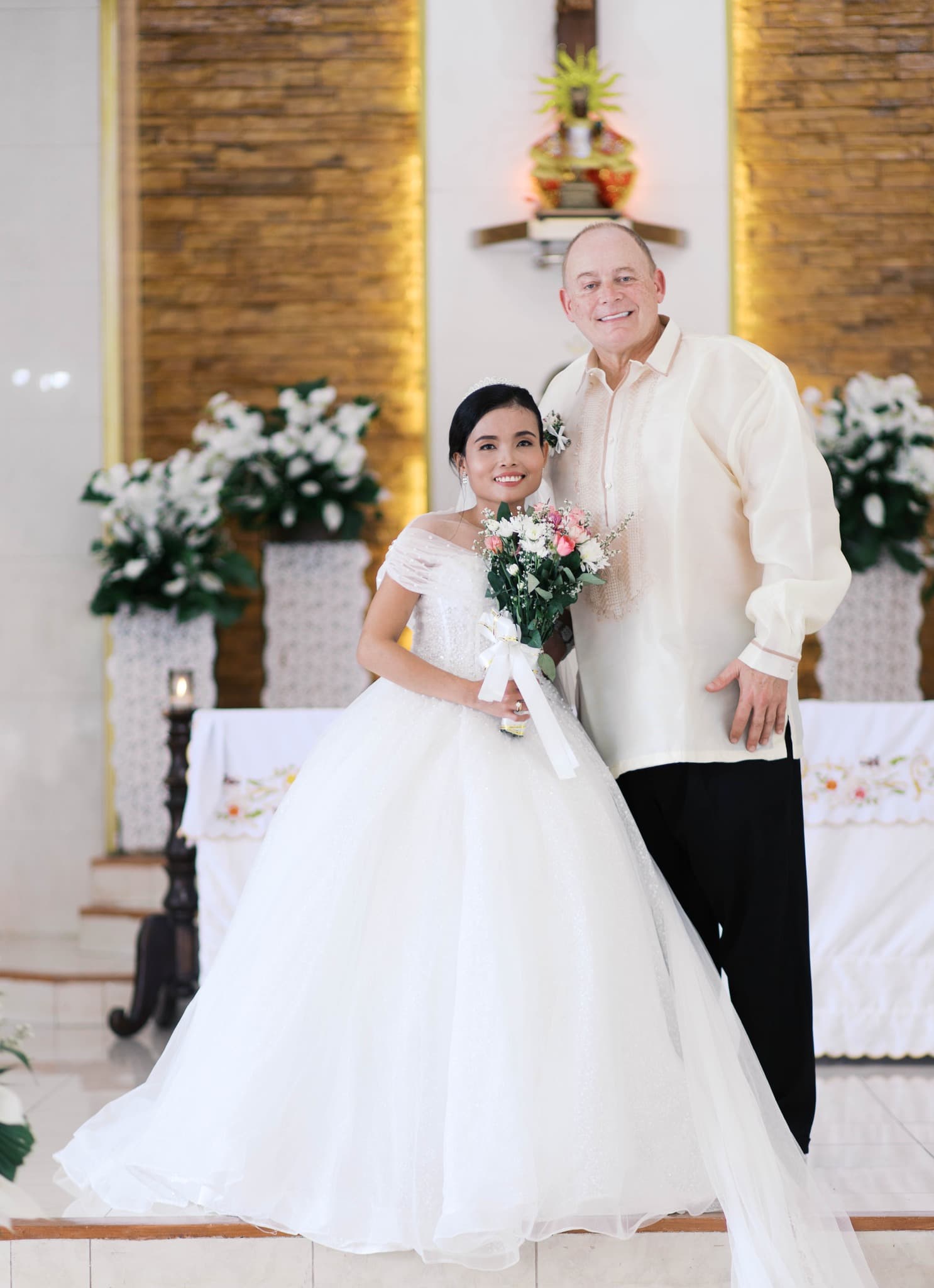 Smiling bride in white gown and groom in Barong Tagalog pose together in a church.