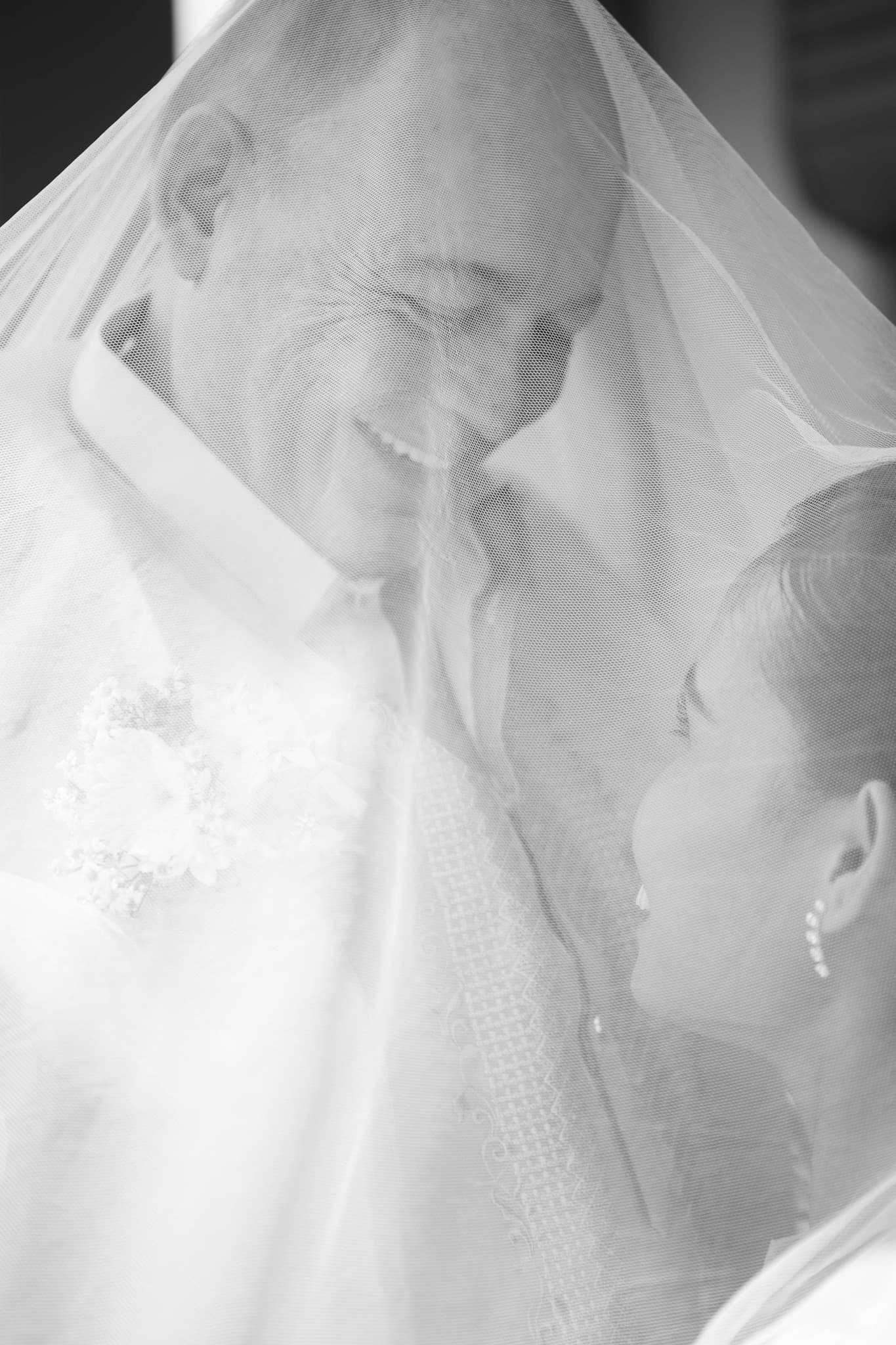 Black and white portrait of a smiling bride and groom under a sheer wedding veil.