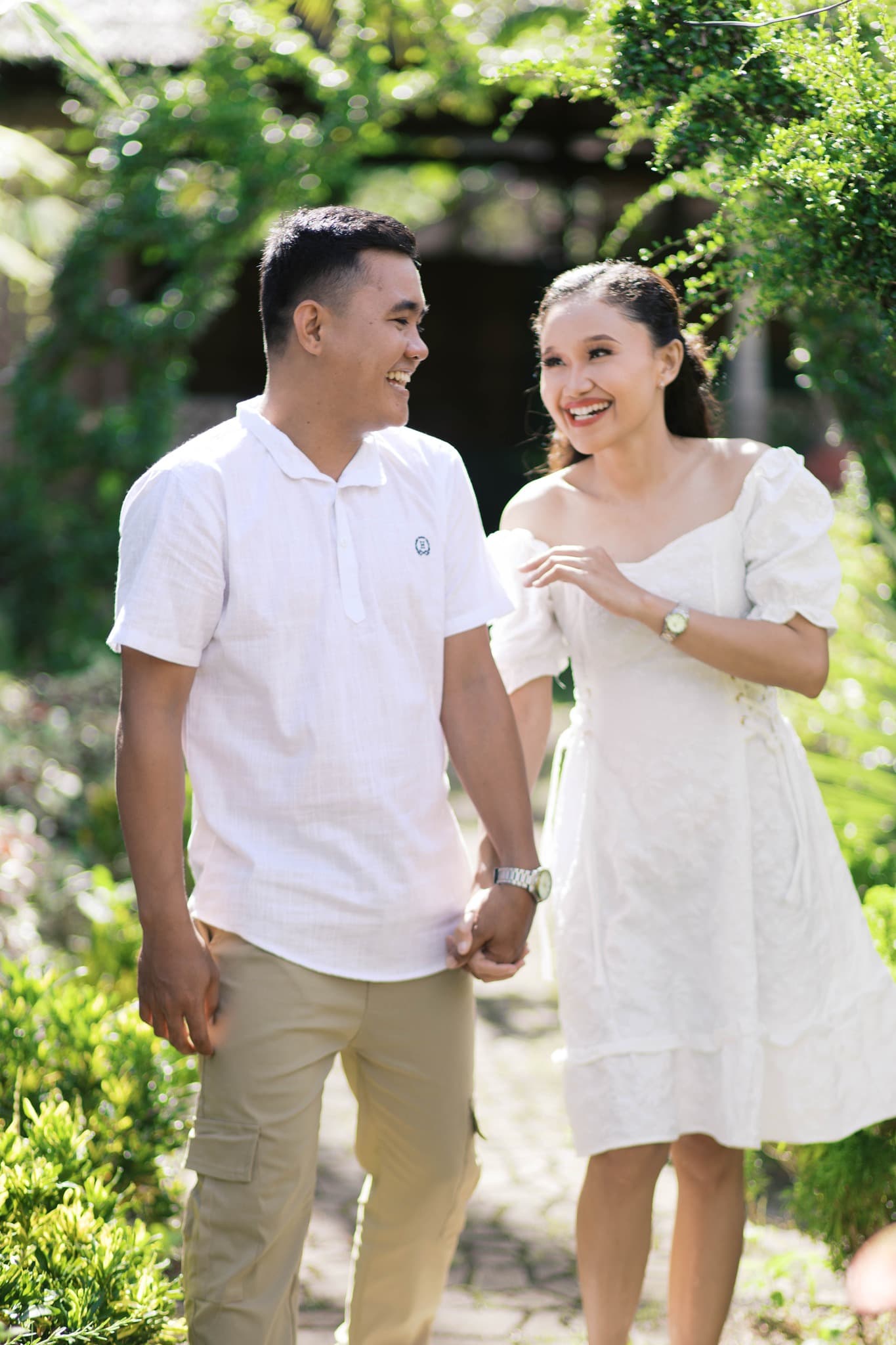 Happy couple in white attire holds hands and laughs while walking through a lush garden.