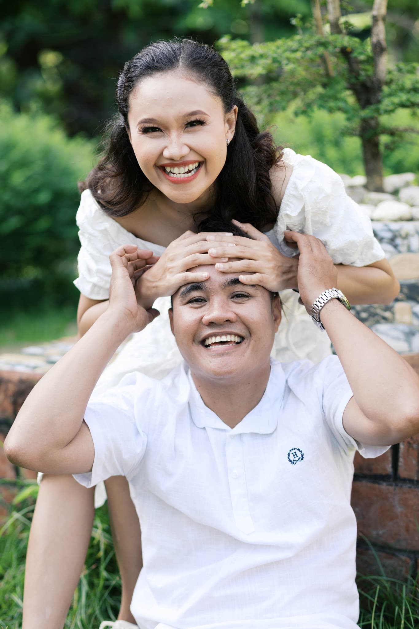 Happy couple in white laughing outdoors, woman resting her hands on the man's forehead.