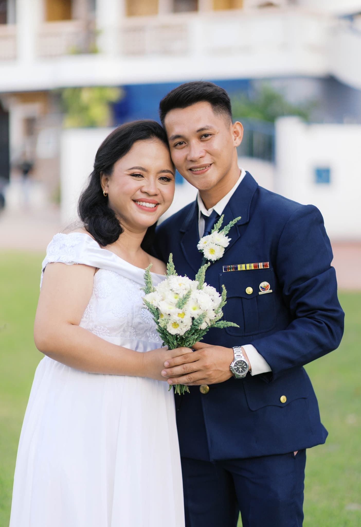 Smiling bride in white dress and groom in navy military uniform holding a white bouquet.