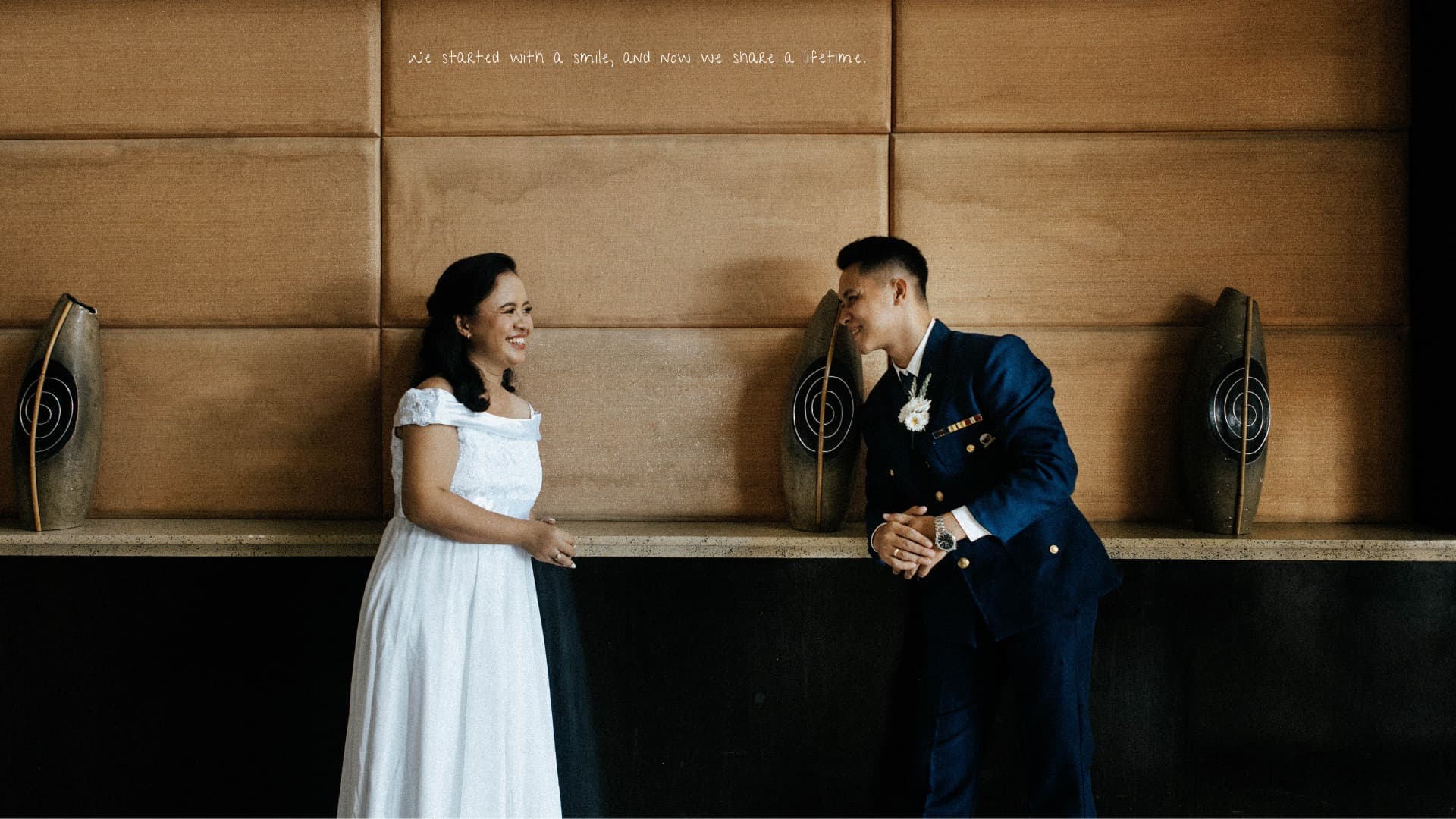 Smiling bride in white and groom in blue uniform against a tan paneled wall.