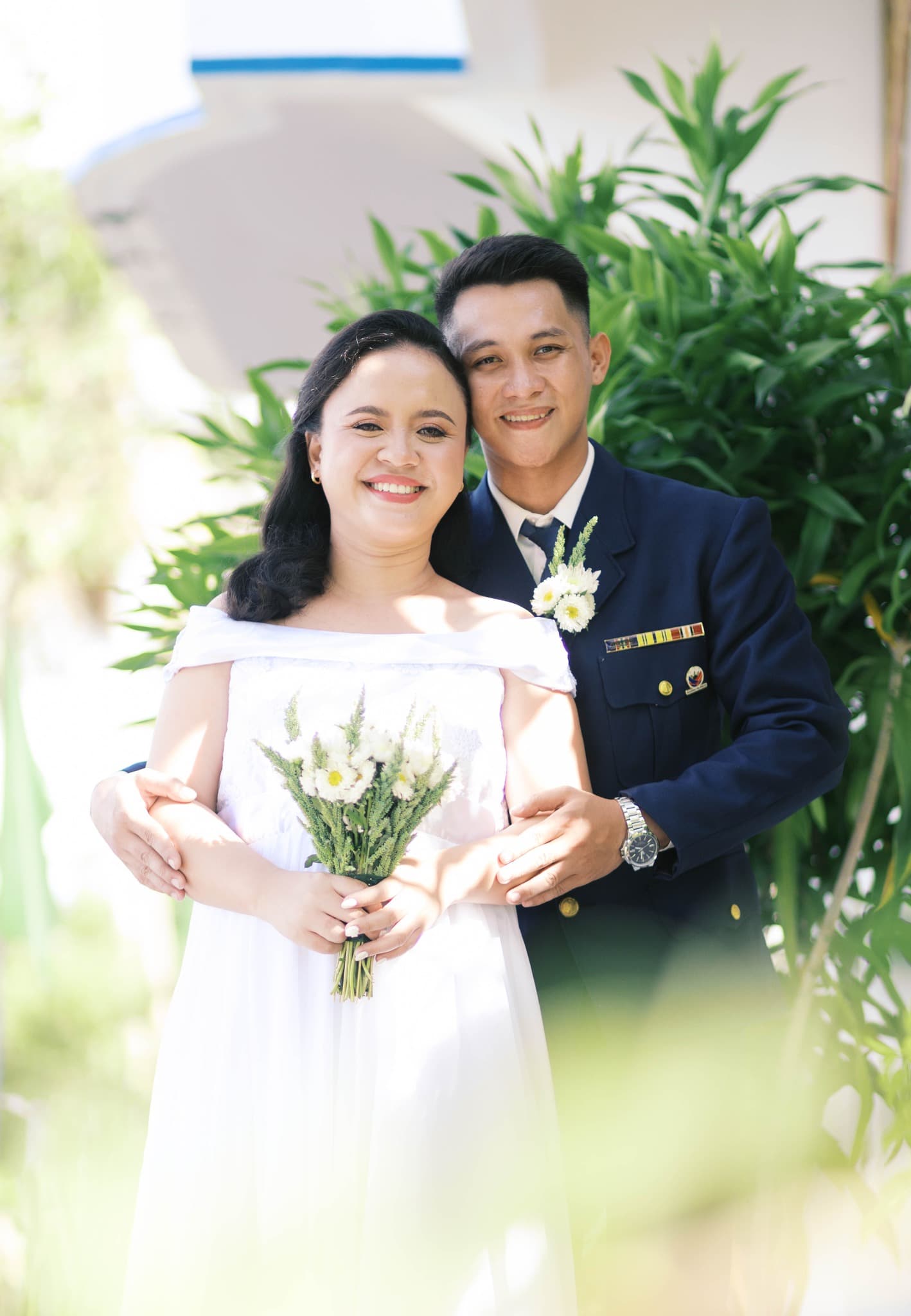 Smiling woman in white dress holding bouquet, man in blue military uniform posing outdoors.
