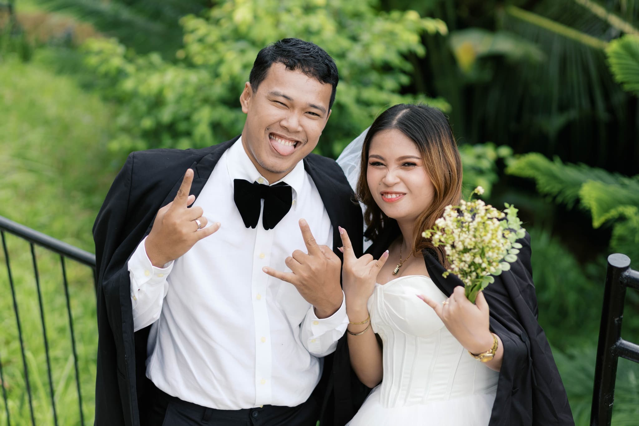 Playful bride and groom making rock-on hand gestures and silly faces in a garden.