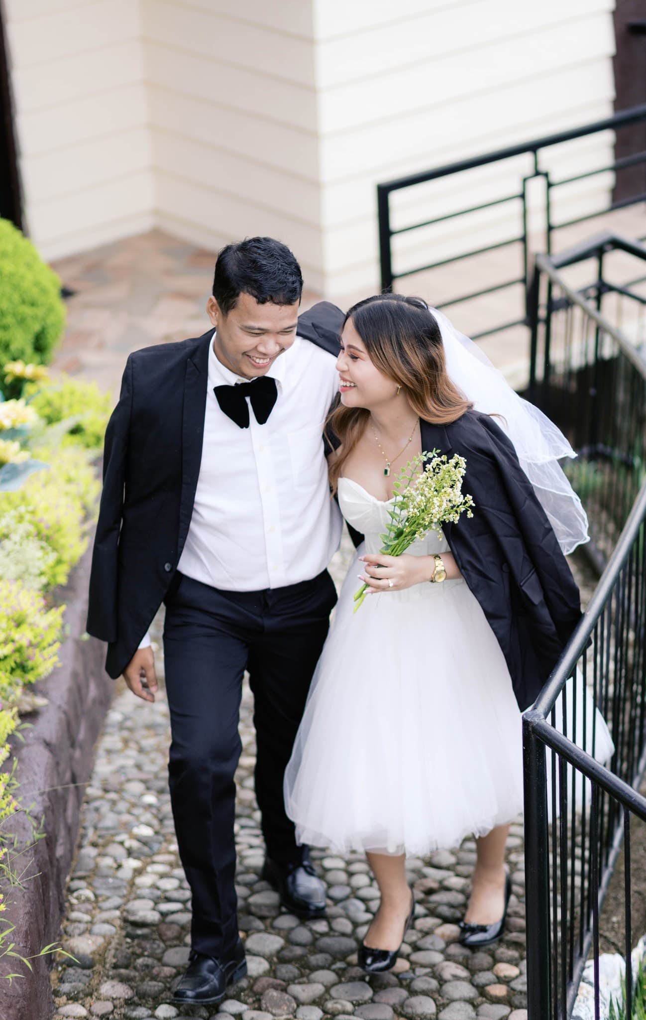 Smiling bride and groom walk down a stone path together, looking at each other happily.