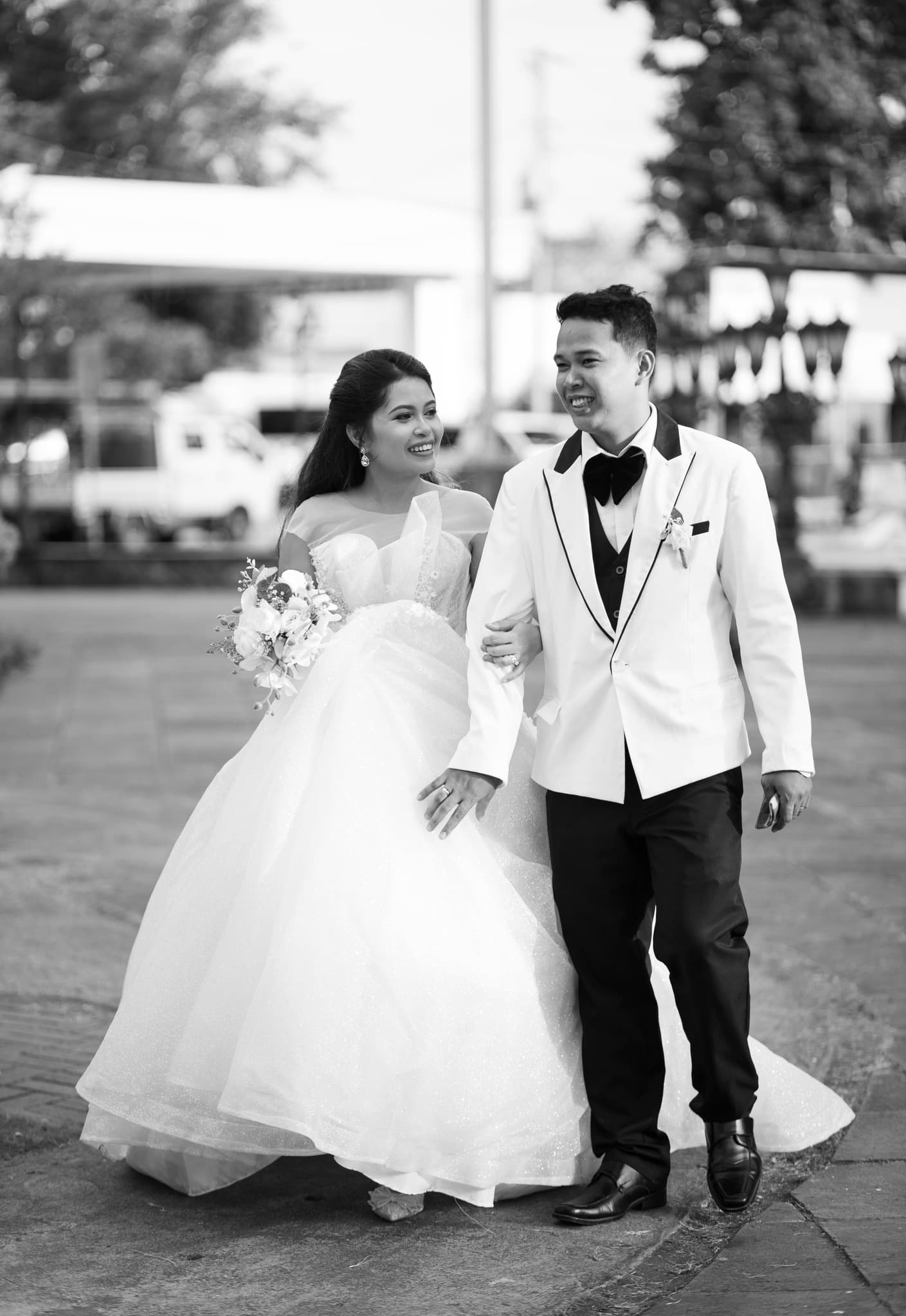 Smiling bride in a white ballgown and groom in a tuxedo walk together outdoors.