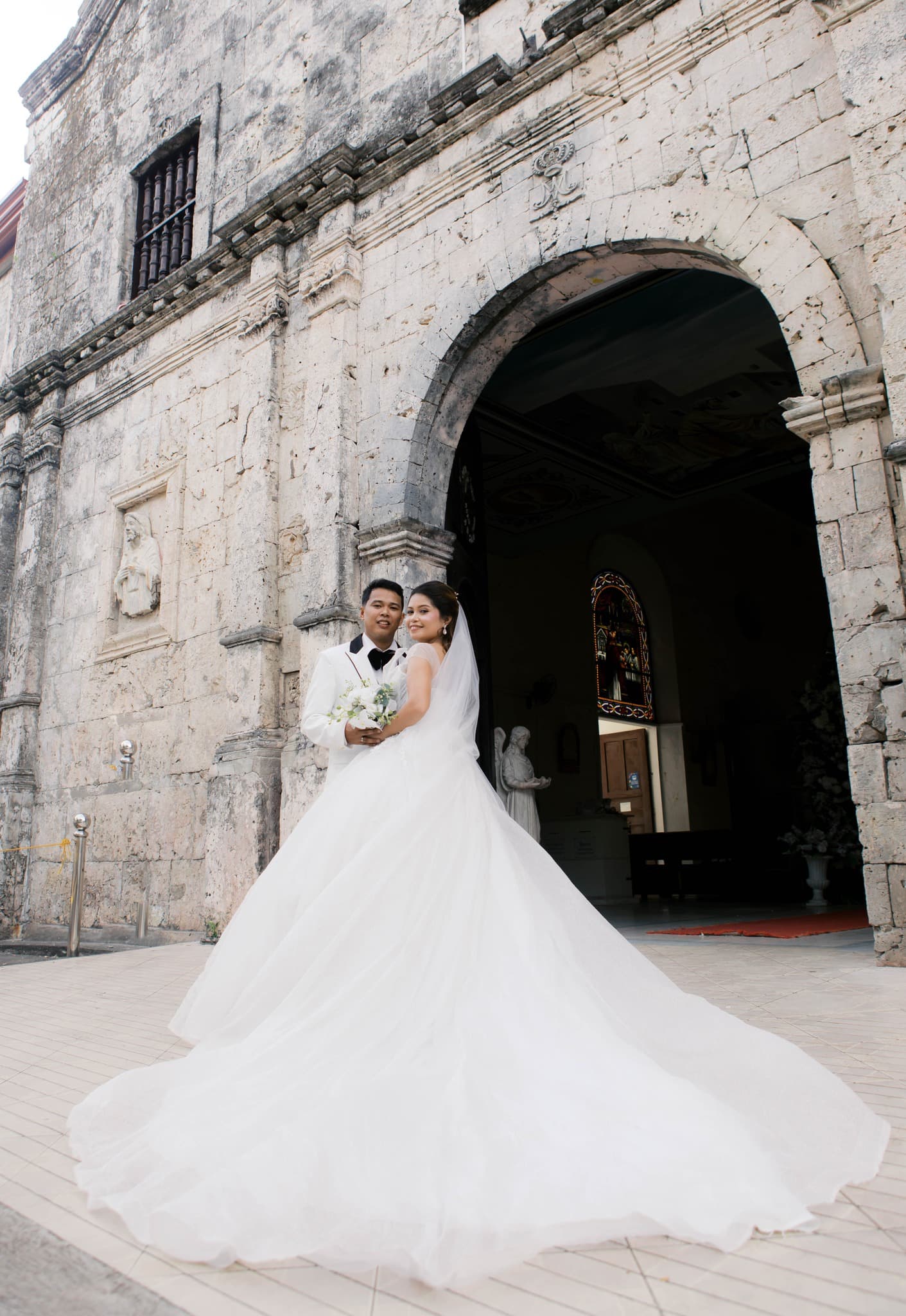 Bride in voluminous white gown and groom in white suit pose before a stone church.