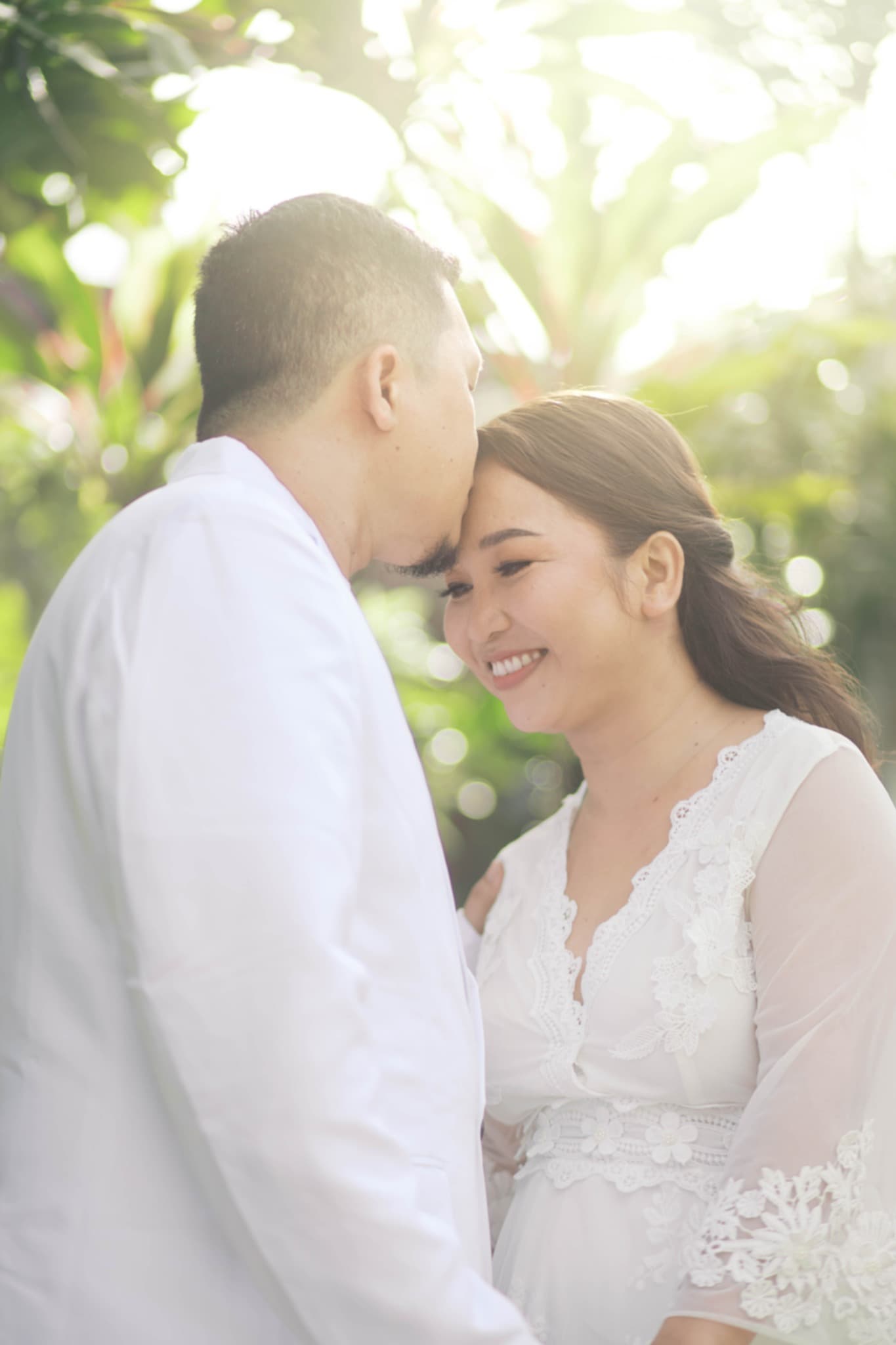 A man in white kisses a smiling woman's forehead amidst bright, sun-drenched outdoor greenery.