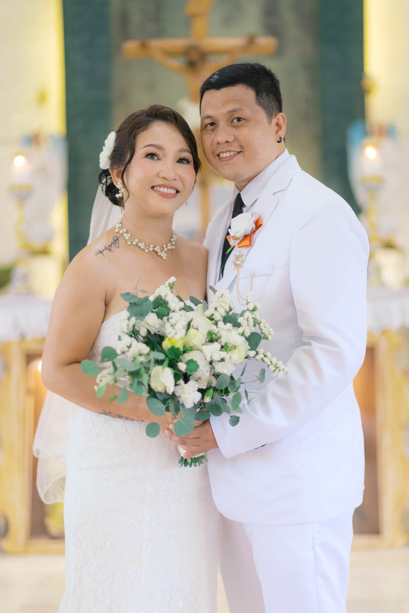 Smiling bride and groom in white wedding attire posing in a church holding a bouquet.