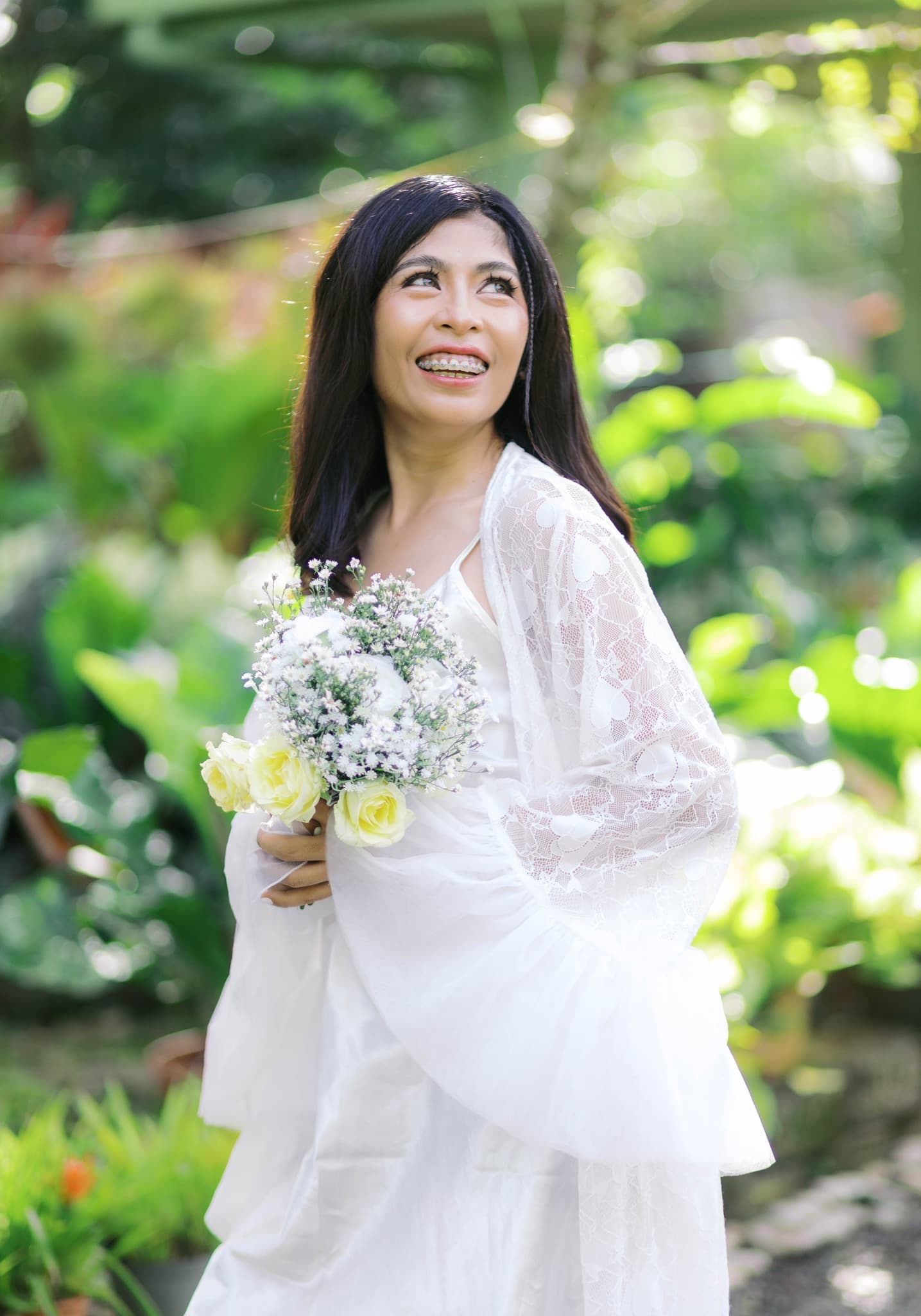 Smiling woman in white lace robe holding a bouquet of flowers in a lush garden.