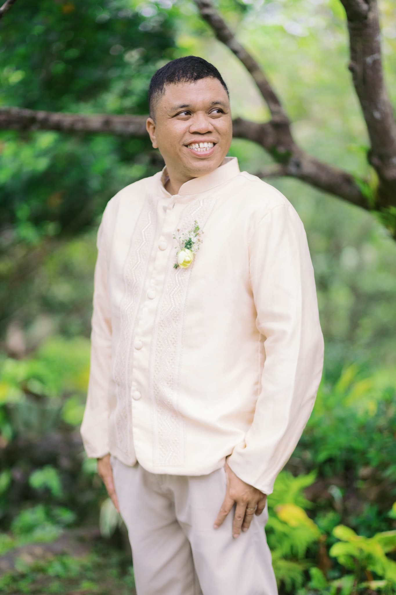 Smiling man in a cream Barong Tagalog with a boutonniere, standing in a garden.