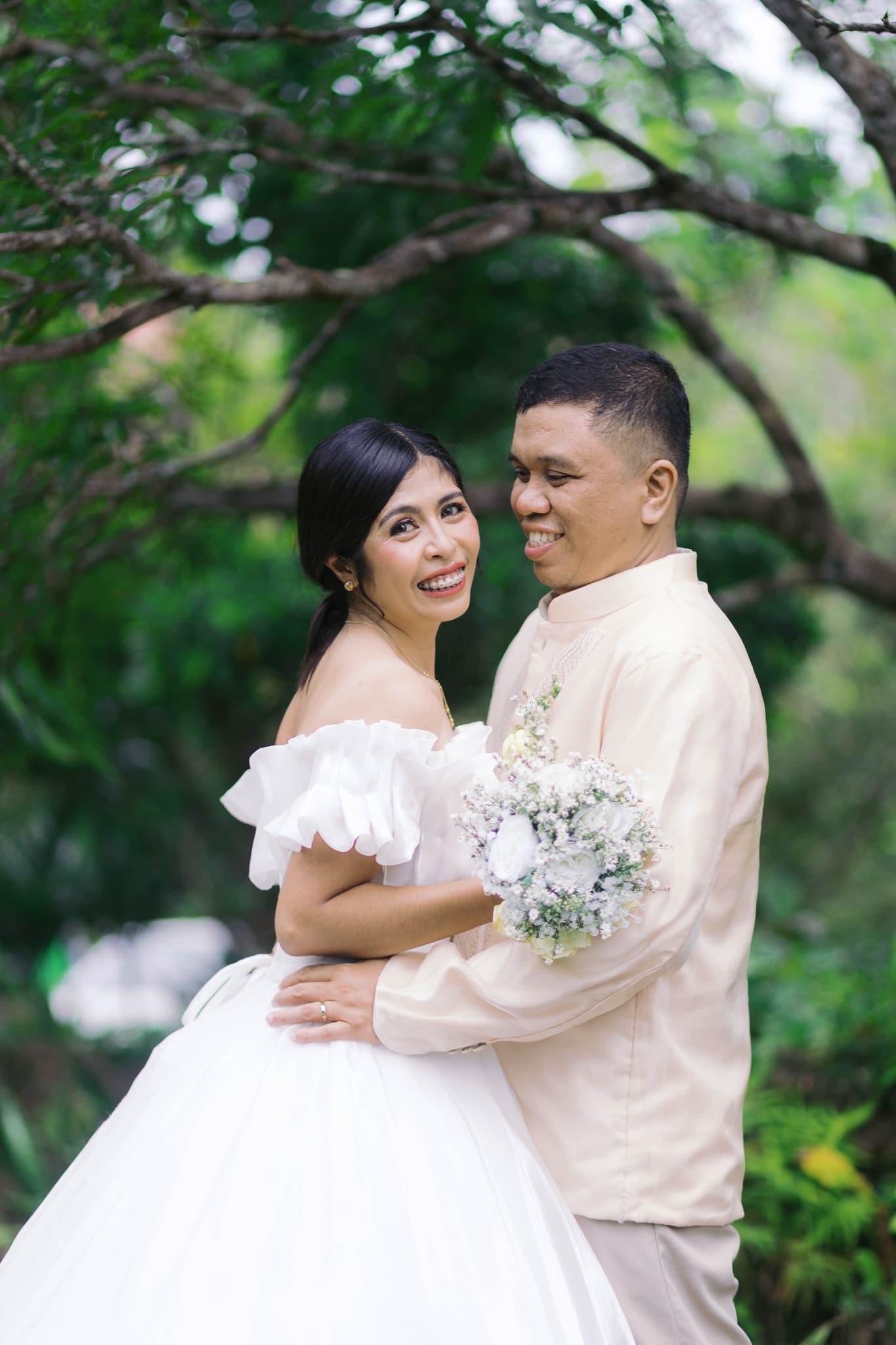 Smiling bride in white gown and groom in Barong Tagalog embrace amidst lush greenery.