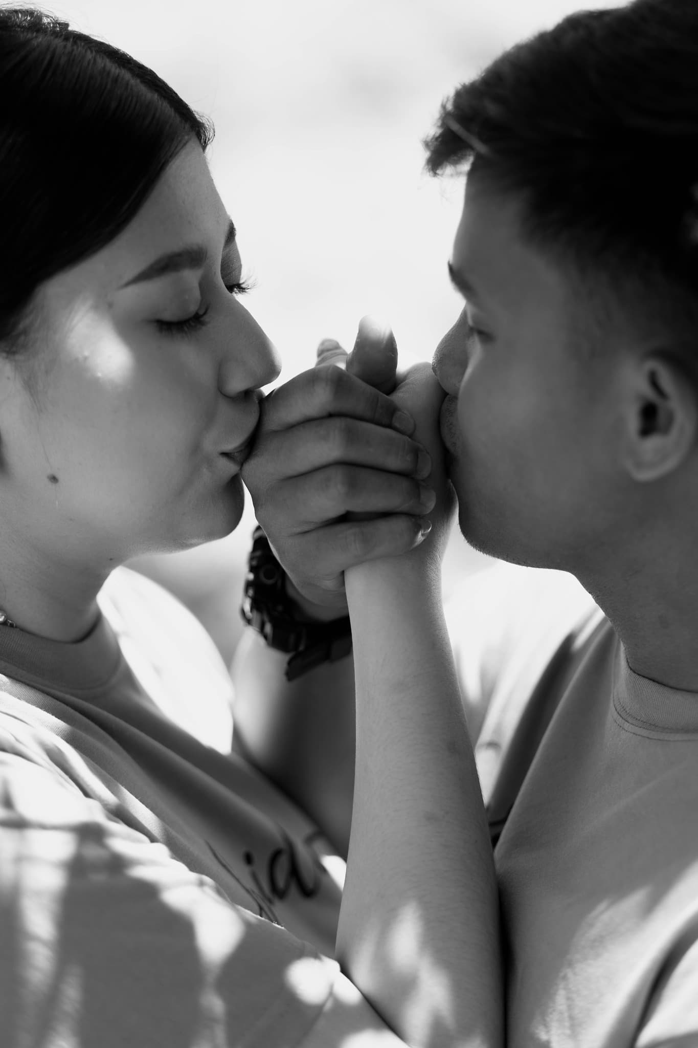 Black and white close-up of a couple tenderly kissing their clasped hands with eyes closed.