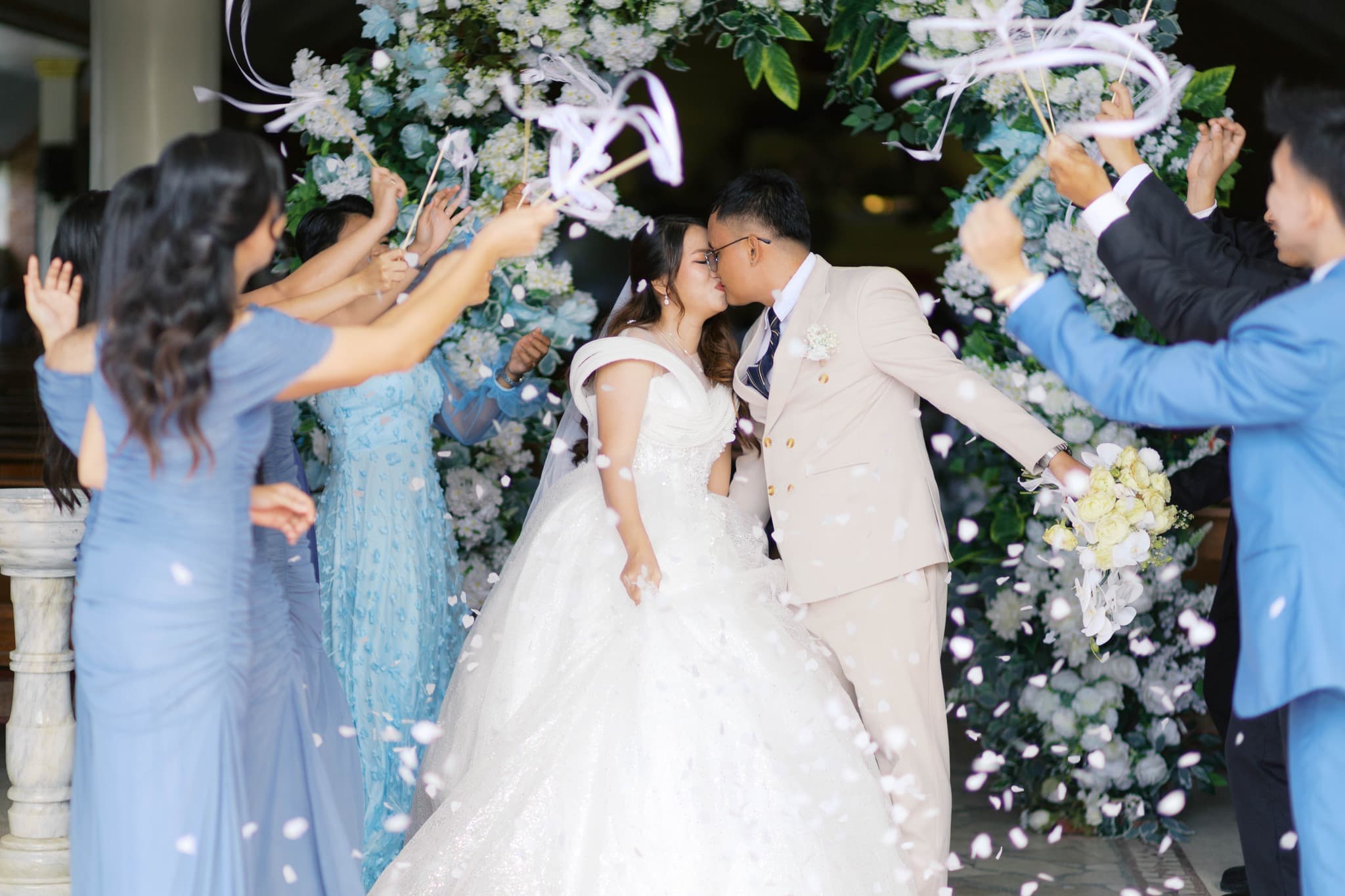 Newlyweds kiss under a floral arch as guests wave ribbon wands and toss white petals.