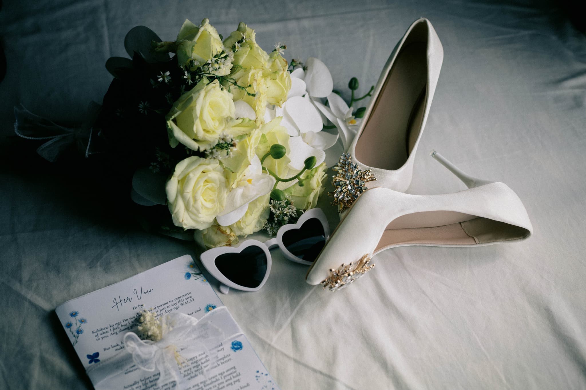 White jeweled heels, yellow rose bouquet, heart-shaped sunglasses, and wedding vows on white fabric.