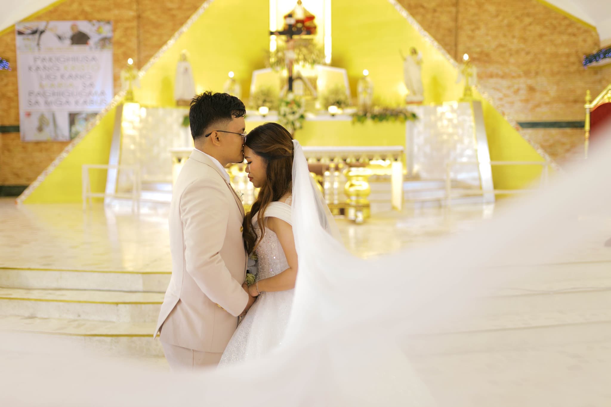 Groom kisses bride's forehead at a golden church altar, her long white veil flowing softly.