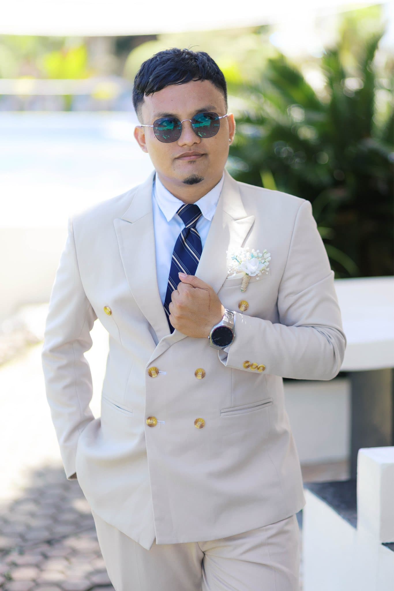 Man in a beige double-breasted suit, sunglasses, and white boutonniere posing outdoors.