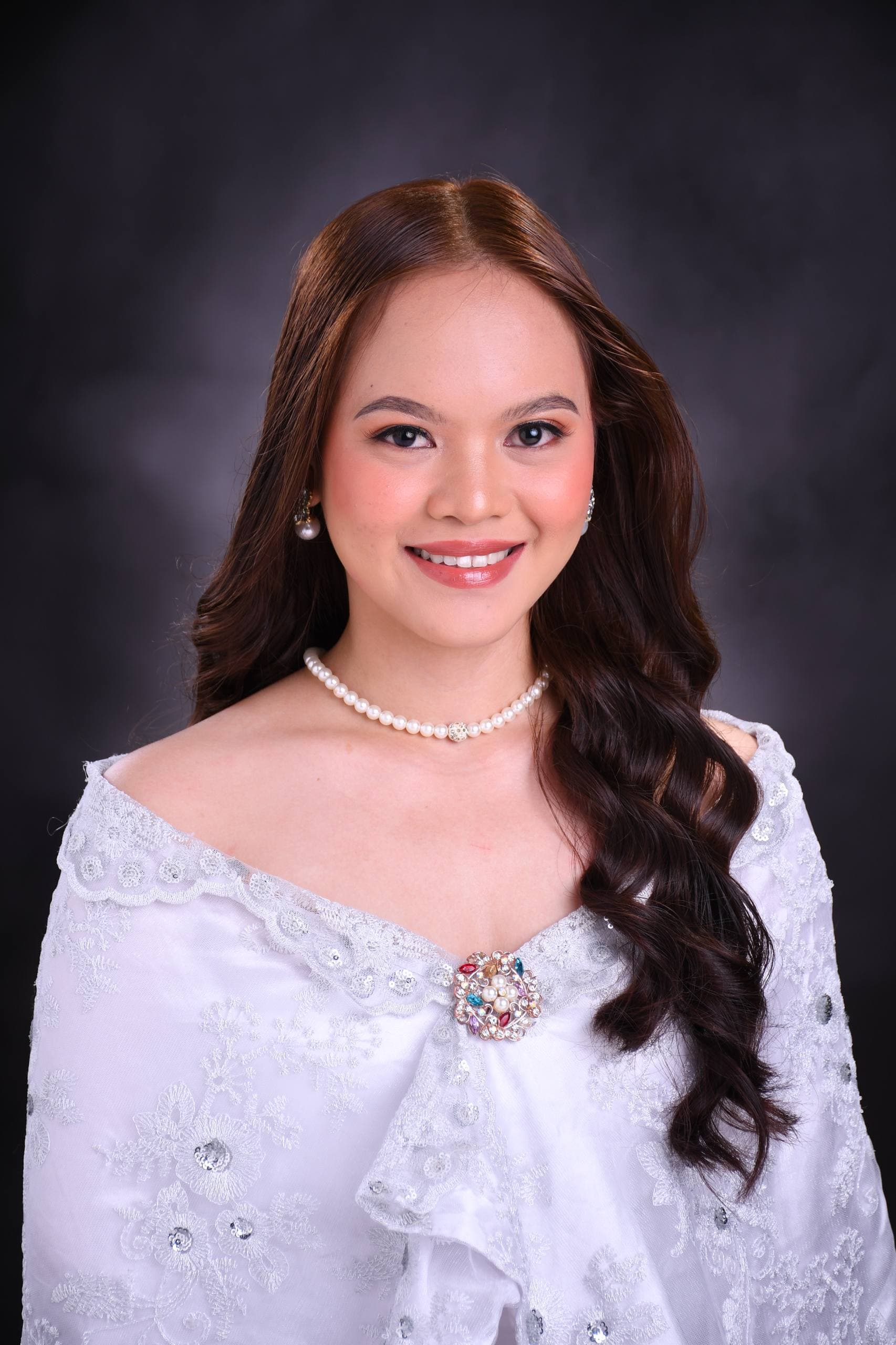 Smiling woman with long brown hair wearing a white lace Filipiniana dress and pearl necklace.